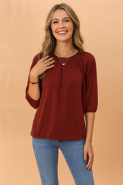 Bordeauxrode dames top met ronde halslijn en driekwartmouwen, afgewerkt met subtiele plooien bij de hals voor een flatterende en elegante uitstraling. Gemaakt van een zachte jersey stof met een losse, comfortabele pasvorm en elastische mouwboorden.