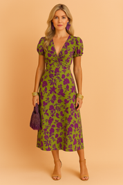 Vrouw draagt een groene midi-jurk met korte mouwen en paarse bloemenprint, met een overslag V-hals en een flatterende taille, tegen een warme beige achtergrond.