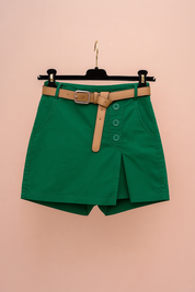Flat-lay van een groene skort met hoge taille, overslagstijl en decoratieve knopen, afgewerkt met een beige riem — minimalistische studio-opname van een zomerse must-have.