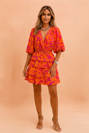 Korte zomerjurk met tropische bloemenprint in oranje en fuchsia, diepe V-hals, ballonmouwen en gelaagde rucherok voor een opvallende feestelijke look.