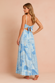 Vrouw draagt een lichtblauwe en witte maxi-jurk met bloemenpatchwork, dunne bandjes en kanten afwerking, en houdt een witte clutch vast tegen een warme perzikkleurige achtergrond.