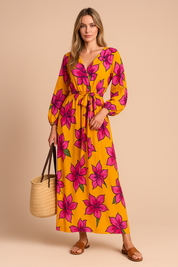 Lange V-halsjurk met strikceintuur en ballonmouwen, bedrukt met grote roze bloemen op een feloranje achtergrond.