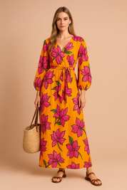 Lange V-halsjurk met strikceintuur en ballonmouwen, bedrukt met grote roze bloemen op een feloranje achtergrond.
