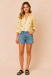 Gele blouse met zwarte zwaluwprint en opgerolde mouwen, gemaakt van ademende linnenmix, gestyled met een denim short.