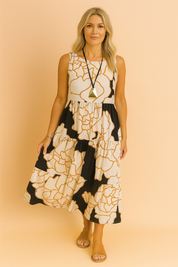 Mouwloze midi-jurk met zwarte en caramelkleurige oversized bloemenprint op een crèmekleurige achtergrond, afgewerkt met ruches in lagen.