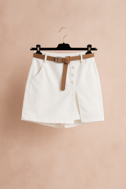 Witte skort met hoge taille, decoratieve knopen en een bruine riem, gestyled met een taupekleurige top en hakken — fris, elegant en modern.