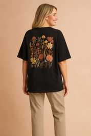 Zwart dames T-shirt met ronde hals en korte mouwen, uitgevoerd in een losse pasvorm. Op de linkerborst bevindt zich een decoratief bloemenmotief in geborduurde stijl, met warme herfsttinten die contrasteren tegen de effen achtergrond. Gemaakt van zachte katoenen jersey, perfect voor casual dagelijks gebruik.