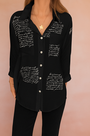 Alt-tekst (NL): Zwarte blouse met handgeschreven witte tekstprint en knoopsluiting aan de voorkant, gemaakt van luchtige stof.