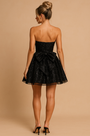 Vrouw draagt zwarte strapless glitter mini jurk met grote strik, achteraanzicht