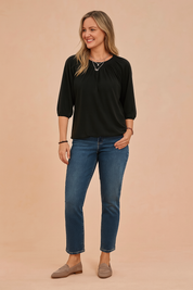 Zwarte dames top met ronde hals en driekwartmouwen, voorzien van elegante plooidetails rond de halslijn en licht gepofte mouwen met elastische boorden. Gemaakt van een soepele, effen jersey stof in een losvallende pasvorm die zowel comfort als een verfijnde uitstraling biedt.